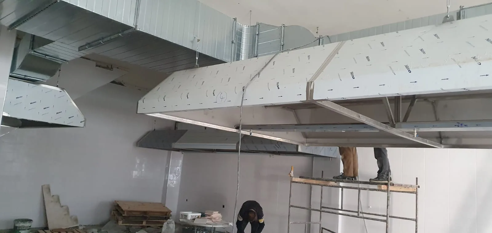 Ticari mutfak hacminde tavana askı sistemiyle asılmış yalıtımlı hava kanalı hattı ve davlumbaz gövdeleri; montaj iskelesi üzerinde çalışan ekip.